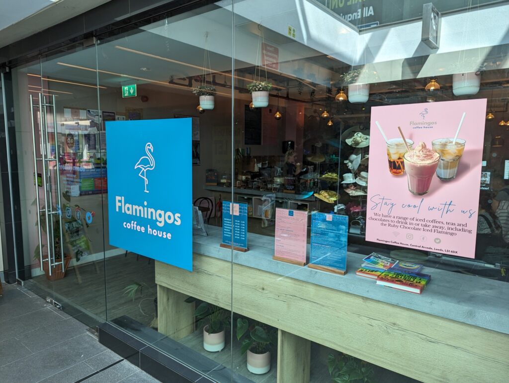 A photo of the exterior of Flamingos Coffee House, showing a glass front with a blue square sign bearing the name of the shop. Wooden tables covered with LGBTQ+ books can be seen inside.